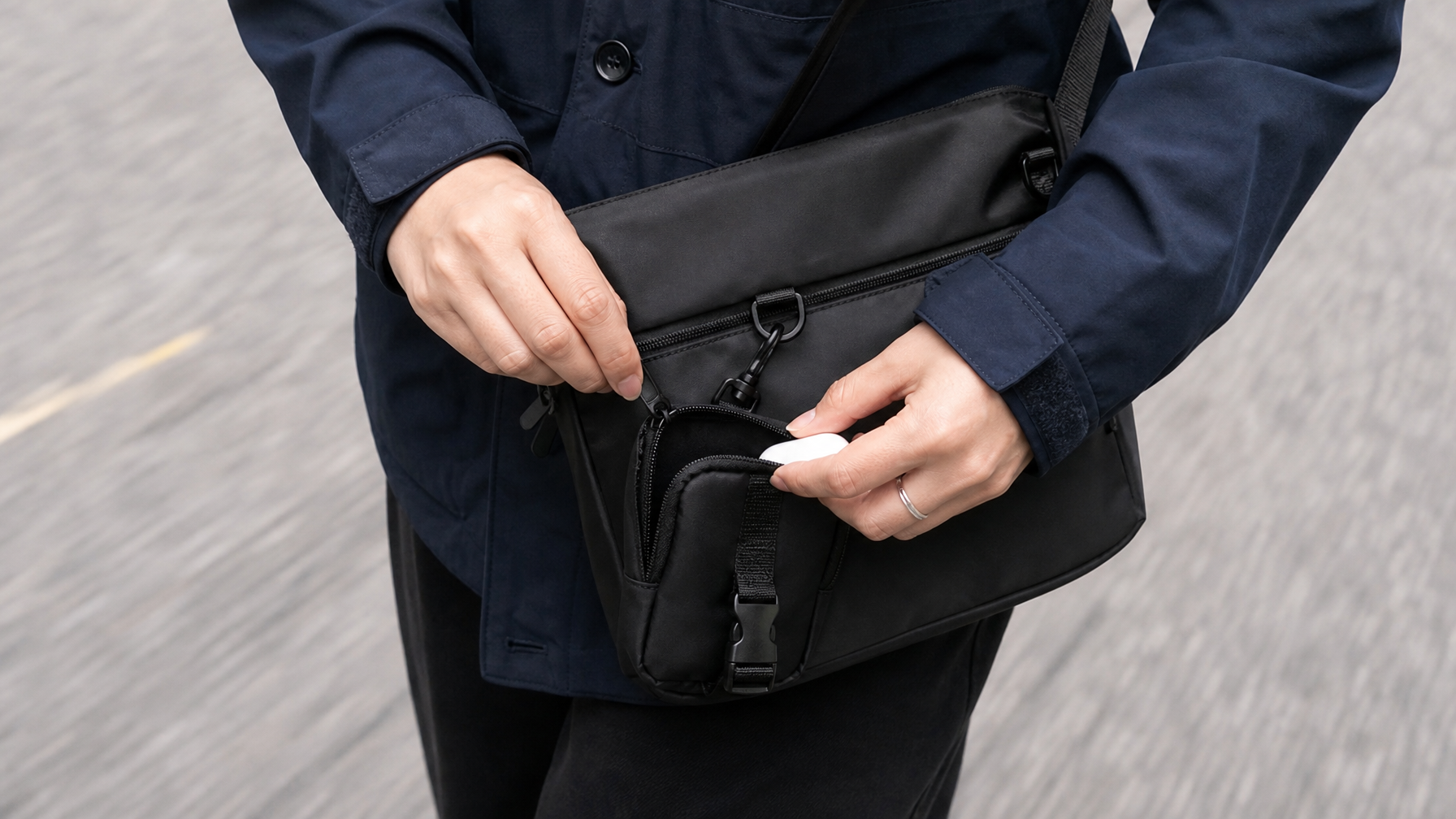 Close-up of a woman's hands opening the front accessory pouch of a black crossbody messenger bag mid-stride on a city street, pavement blurred below