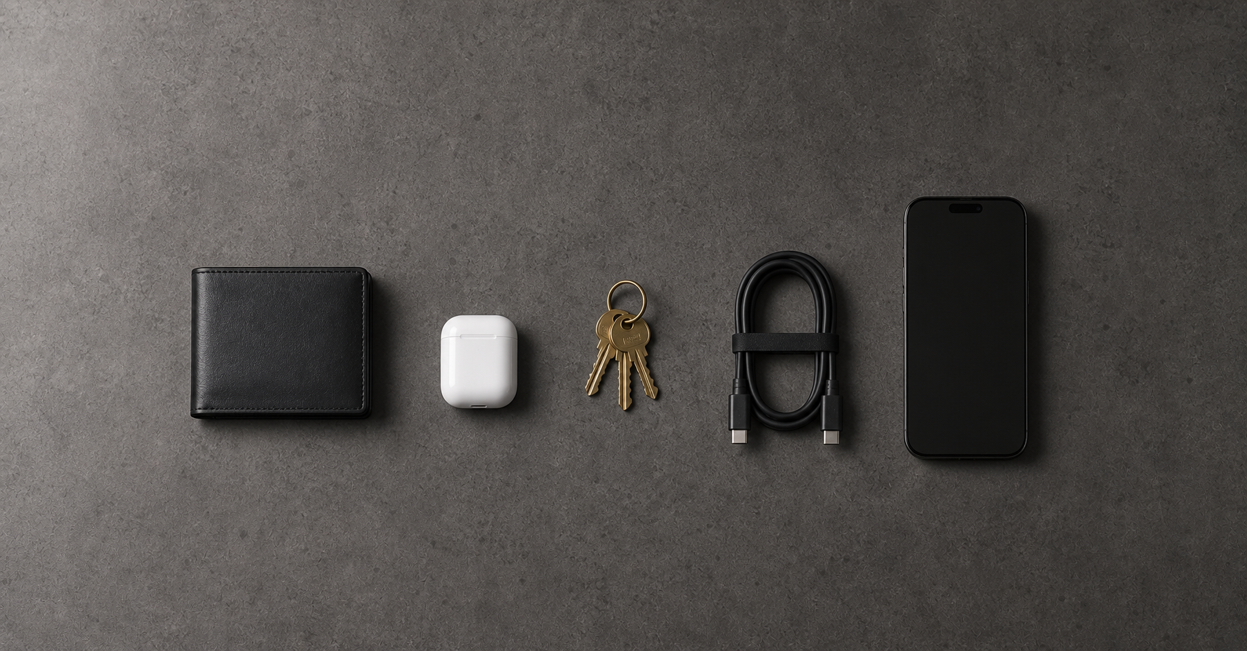 Minimalist EDC essentials flat lay on dark slate concrete — slim wallet, AirPods case, keys, USB-C cable, and phone arranged with intentional spacing in side light