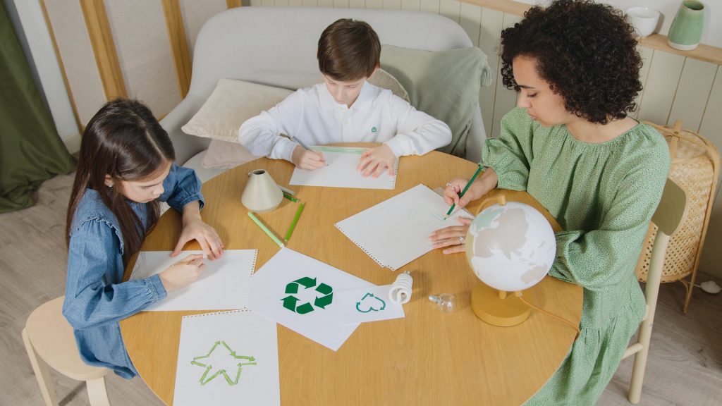Educação ambiental. Na mesa, duas crianças e uma mulher adulta estão desenhando figuras relacionadas á reciclagem e sustentabilidade.