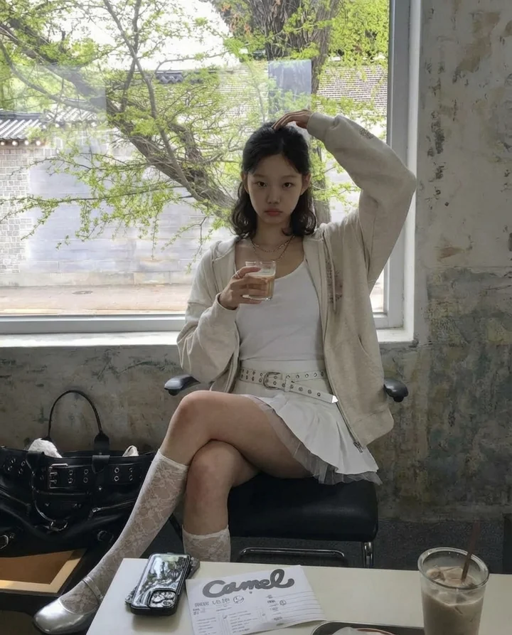 Model Korea berpakaian zip-up krim dan skirt berlipat duduk di kafe cahaya semula jadi, Seoul