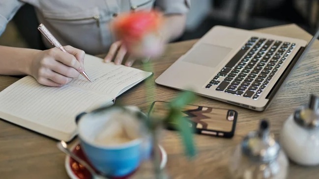 Content writer đang làm việc trên máy tính xách tay, ghi chú ý tưởng bài viết trong sổ tay.  Freelancer viết content, freelancer viết bài, viết lách, dịch vụ viết content, làm content, tuyển dụng content writer.