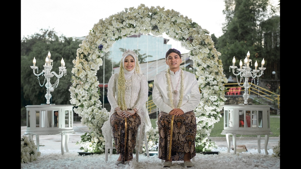 Jasa fotografi prewedding profesional, pasangan pengantin memakai pakaian adat dengan latar belakang dekorasi bunga putih.