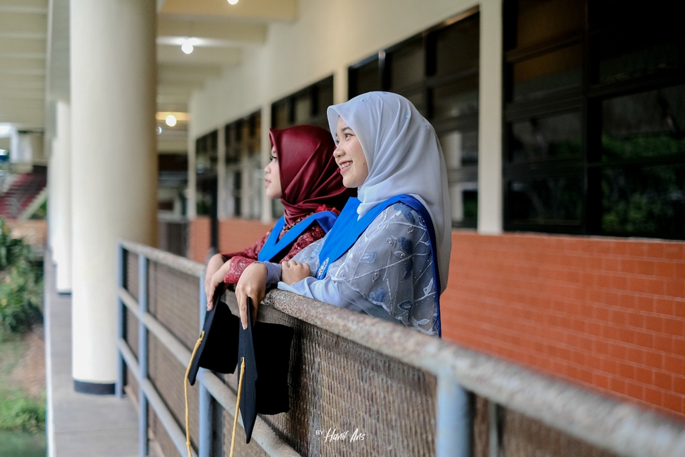 Dua wanita berhijab mengenakan toga wisuda sedang tersenyum dan berfoto di depan balkon. Jasa Fotografi Wisuda, Harga Fotografer Wisuda, Fotografer Wisuda Murah, Jasa Foto Wisuda Outdoor, Sewa Fotografer Wisuda.