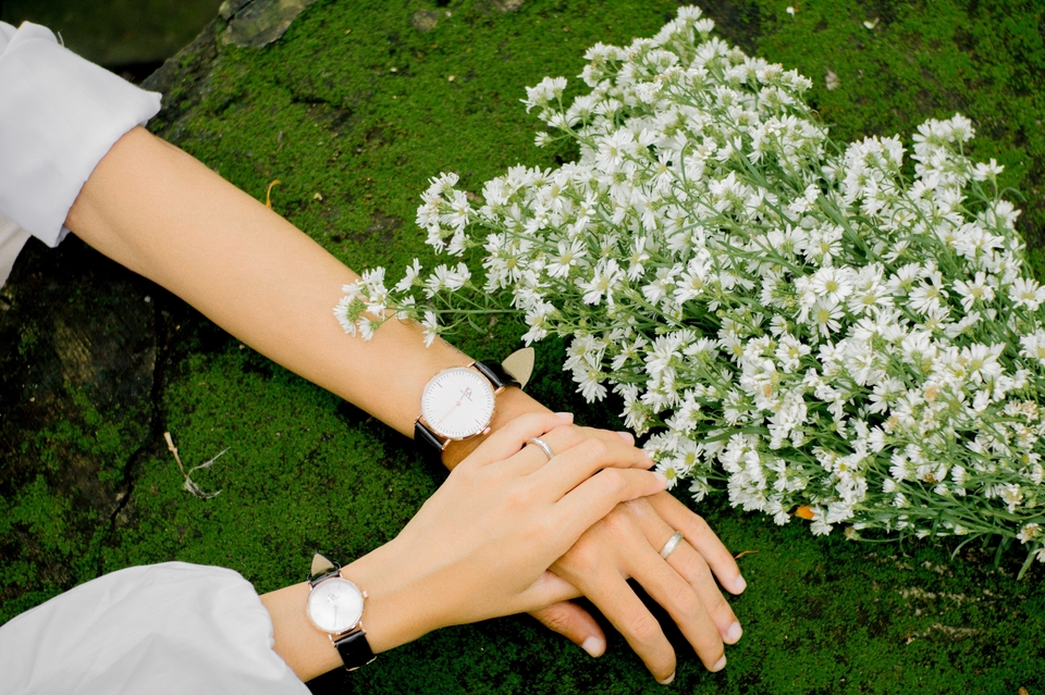 Jasa fotografi prewedding, pasangan memakai jam tangan, latar belakang bunga putih.