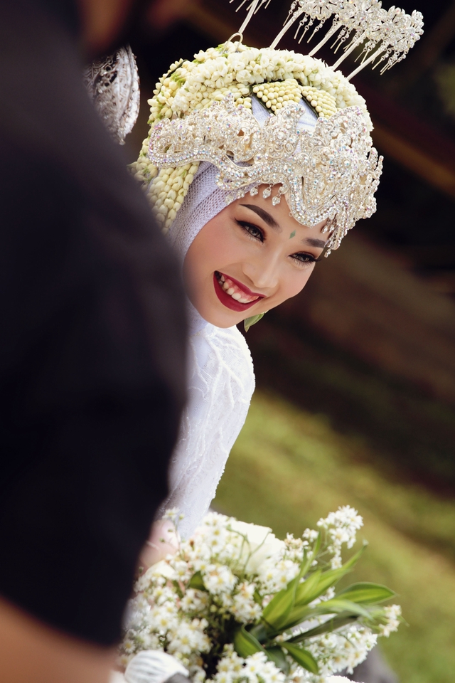 Jasa Fotografi Pernikahan, Wanita cantik berhijab tersenyum di depan kamera dengan bunga putih dan hiasan kepala tradisional