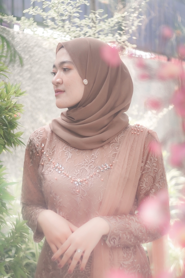 Jasa fotografi prewedding dengan model wanita berhijab mengenakan kebaya dengan desain bunga  di sebuah taman dengan background  pemandangan hijau dan bunga berwarna pink.