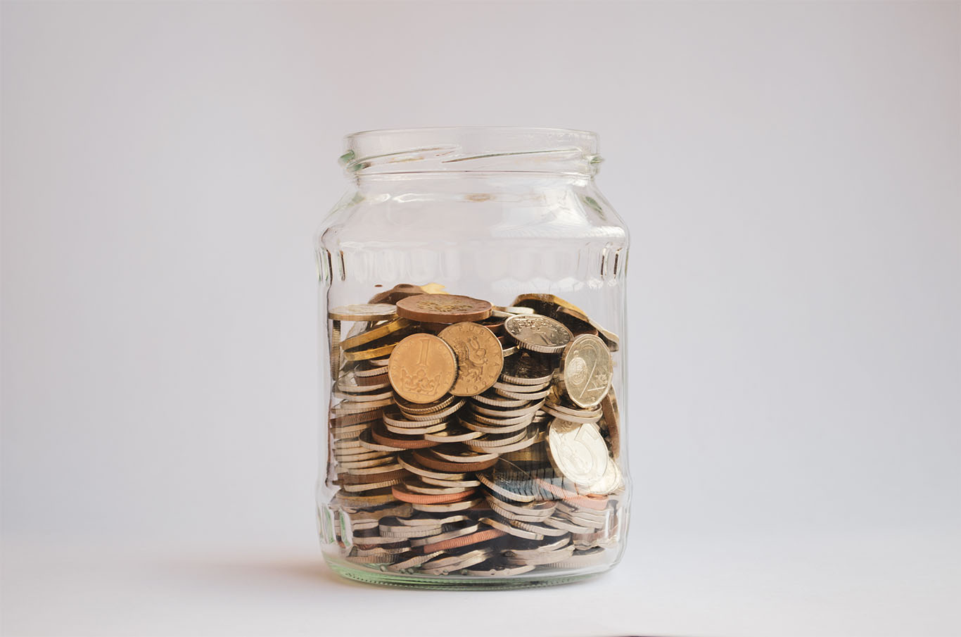 Sklenice plná mincí různých měn na bílém pozadí, symbolizující spoření a finanční úspory.