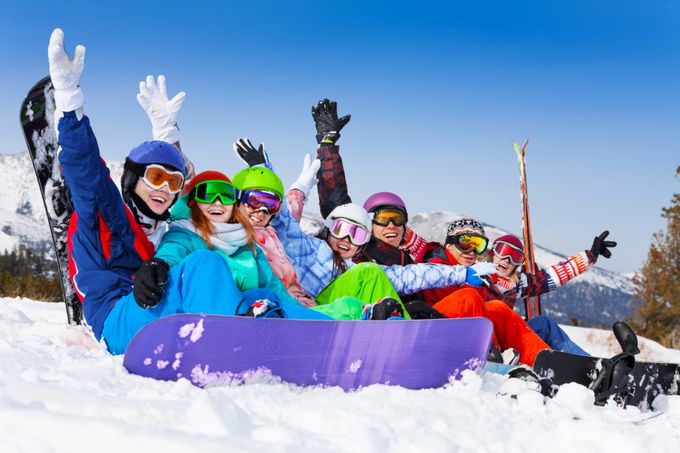 Seven happy smiling friends wearing ski mask sitting together lifting hands up in the air on the mountains background