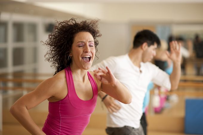 A multi-ethnic group of men and women in a zumba dance fitness class in an indoor gym studio, standing in a row, wearing exercise clothing, and moving to the music.