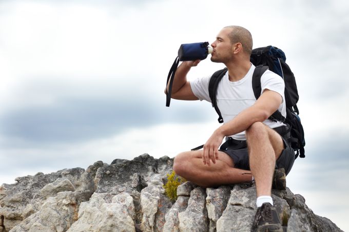 A young man taking a break from hiking while sitting on a rock and drinking some water