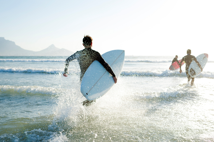 Three teenage boys wearing their wetsuits carrying their surfboards run into the sea on a sunny Summers day.