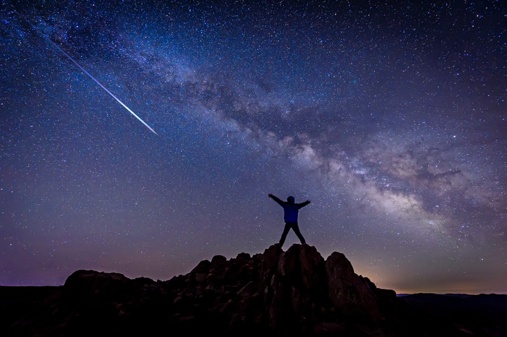 Long exposure image including: bright shooting star (pressed, meteor) next to the beautiful Milky Way Galaxy and countless stars on a clear summer starry night as seen from asia's Northern hemisphere. Silhouettes of man and hill as foreground to frame the shot. Light pollution evident on the horizon from nearby towns and human settlements. Shot on Nikkor D4s,lens by 14-24.