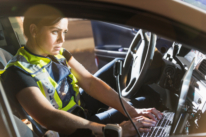 A female police officer sitting in her police car, using the computer. It is nighttime and the emergency lights on her vehicle are flashing.