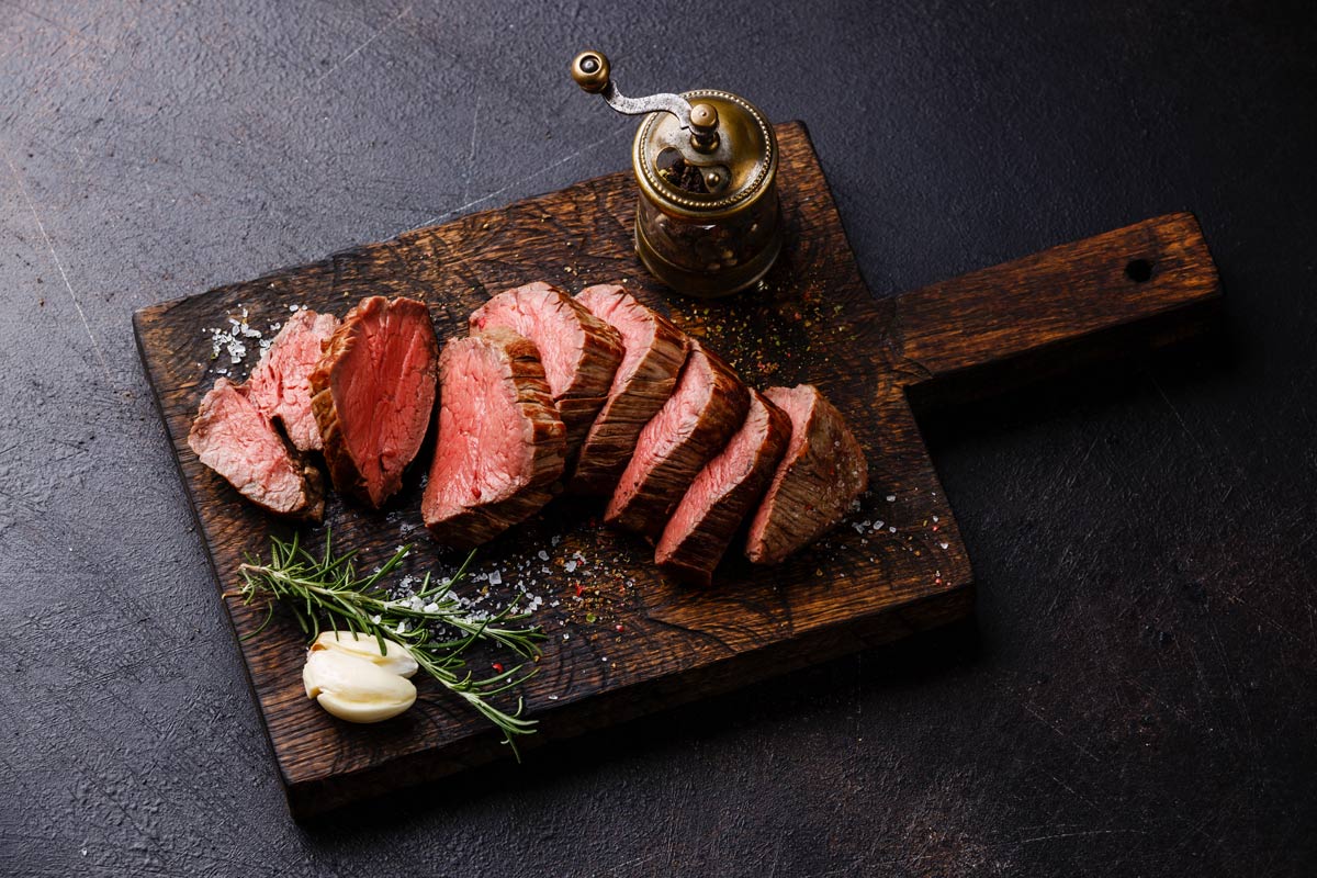 Slices of medium rare roast beef meat on wooden cutting board, pepper and  rosemary branches on wooden background. Top view. Gourmet food. Raw meat  beef steak. Stock Photo