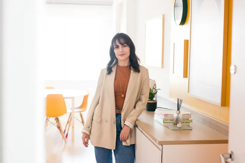 A designer de interiores Jéssica Filla posa para a foto. Ela está apoiada em um balcão e ao fundo há uma mesa redonda com cadeiras amarelas. Vale a pena investir em alugado?