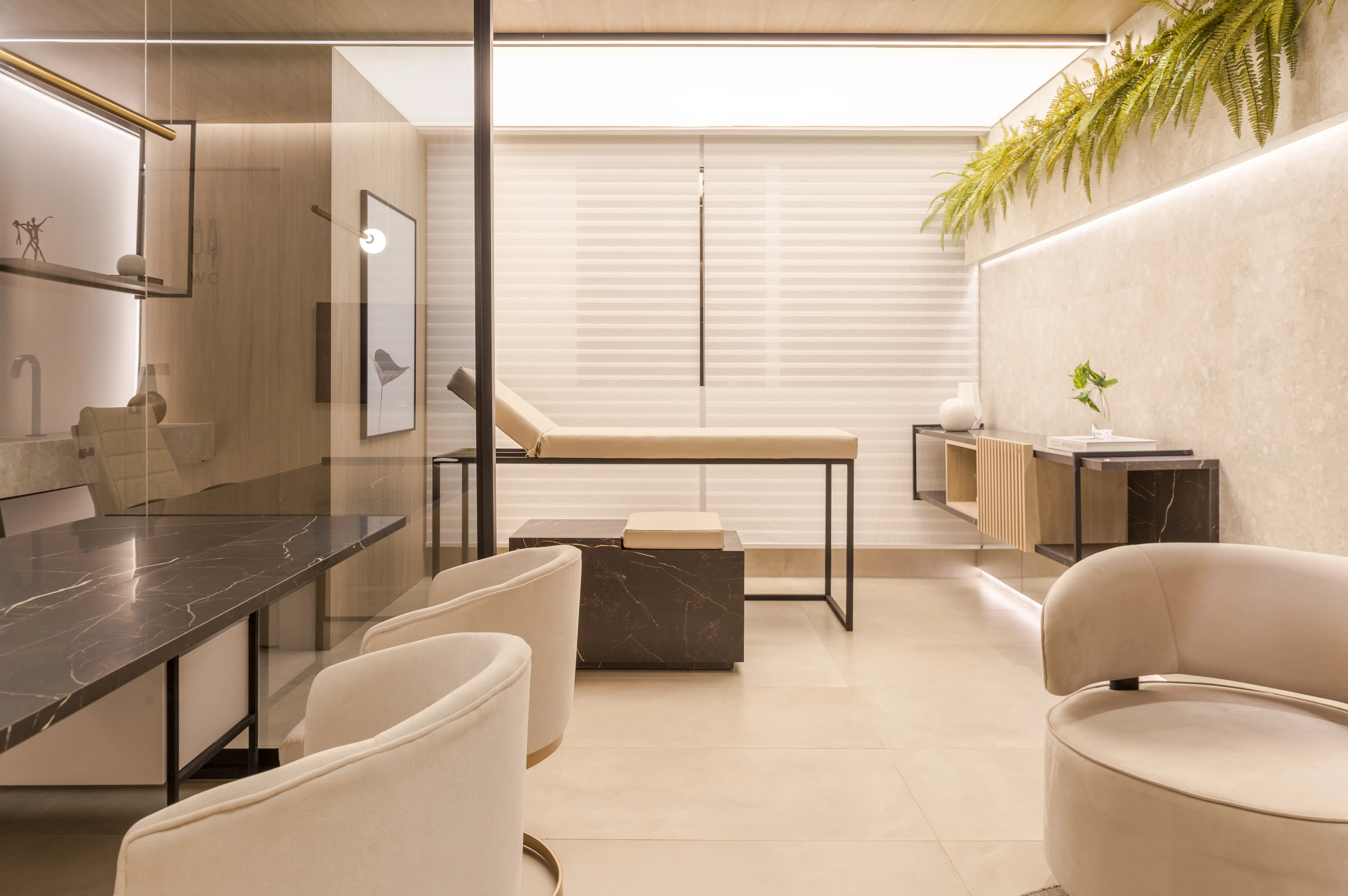 Na imagem, é possível ver a maca, as poltronas do atendimento e a decoração do ambiente - que possui um vidro separador entre a área de atendimento e exame, cores claras e vegetação. 