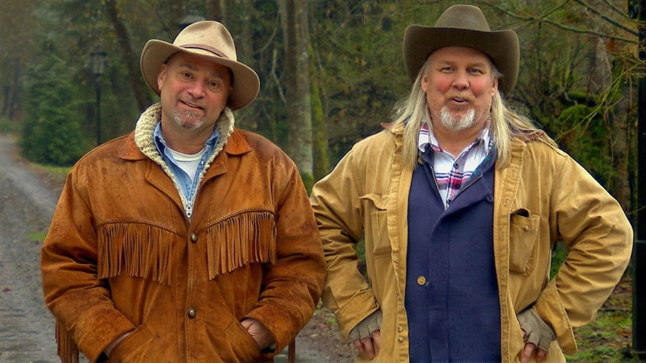 Two men in their middle ages with cowboys hats stood by trees on a dirt path