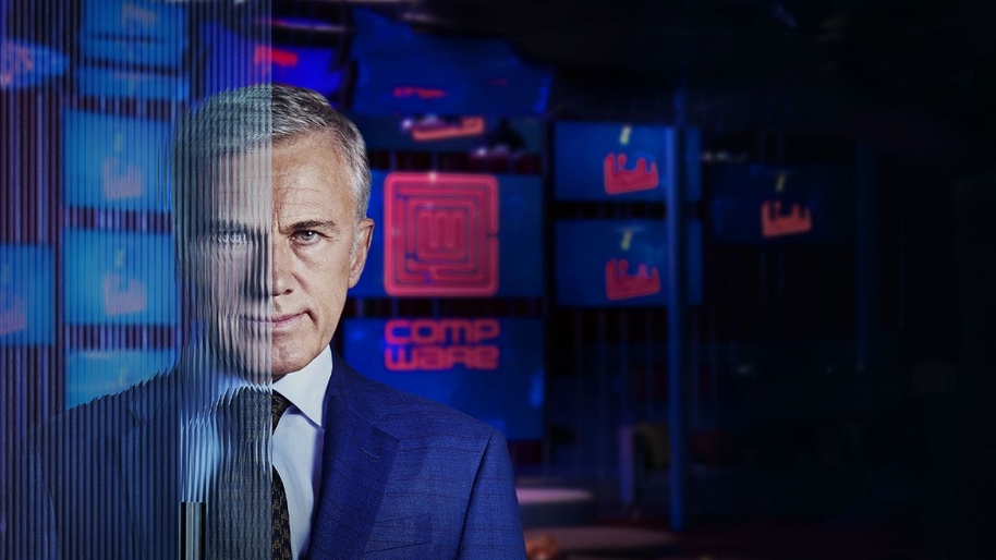A man in a blue suit stood in with screens in the background with half his face behind a screen