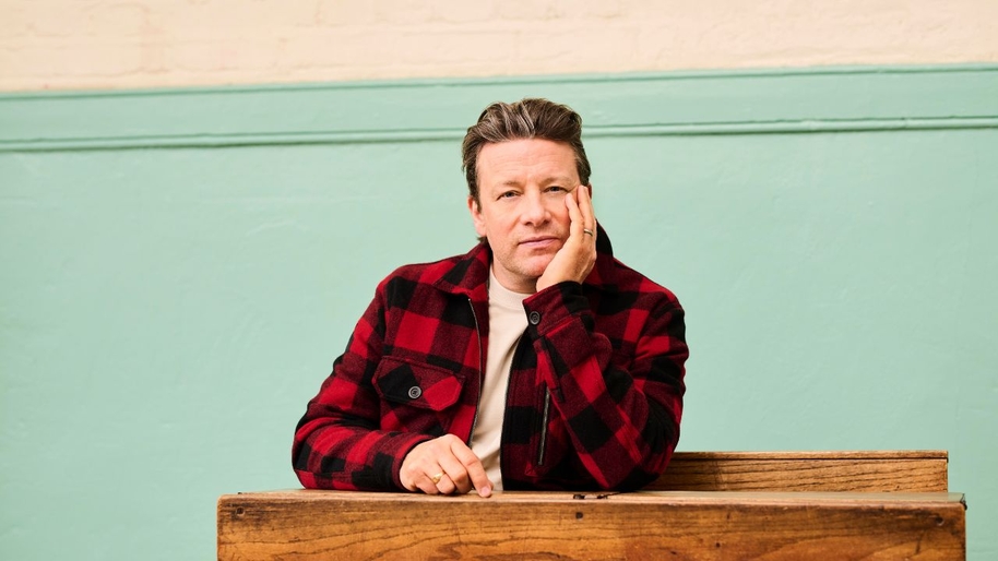 A middle aged man in a red checked shirt sat at a school desk with his left hand against his face