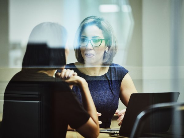 Two business women sitting and talking behind a glass window in the middle of an energetic conversation. One woman has a laptop in front of her