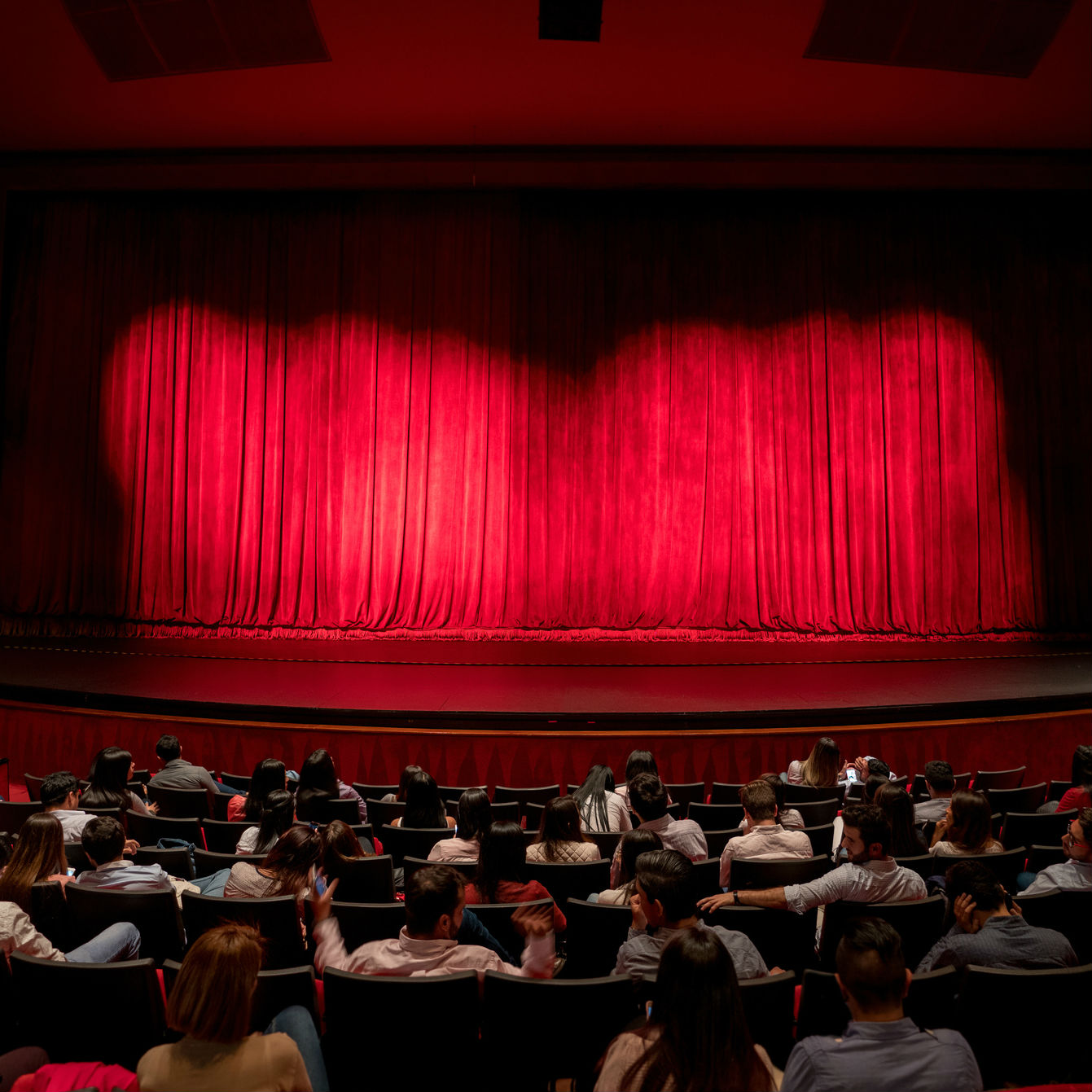 People sitting the theatre seats of a stage with the red curtain closed
