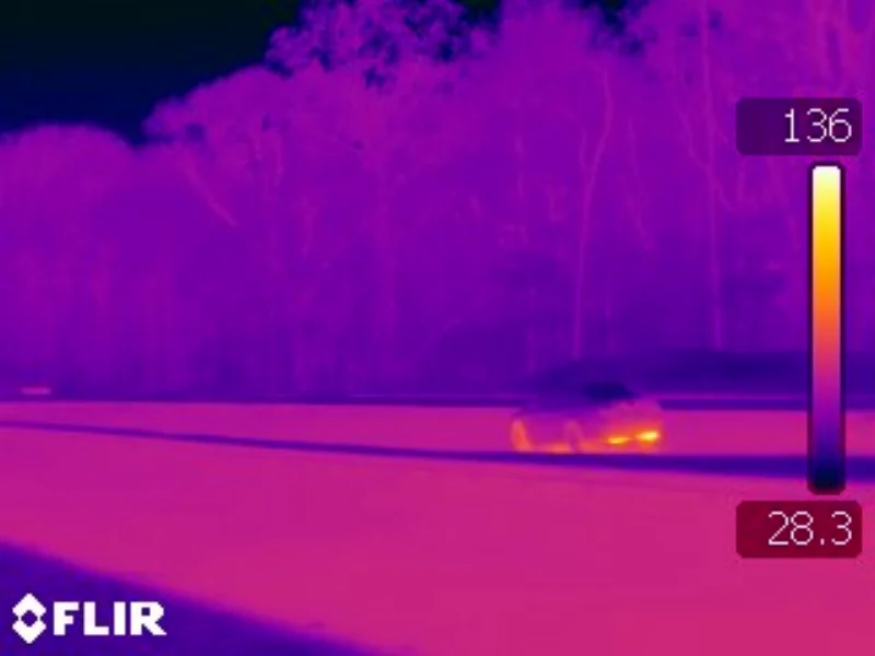 Thermal infrared photograph of a vehicle on a road at night, false-colour heat map showing warm engine and tyre areas against a cool road surface, FLIR temperature scale at right.