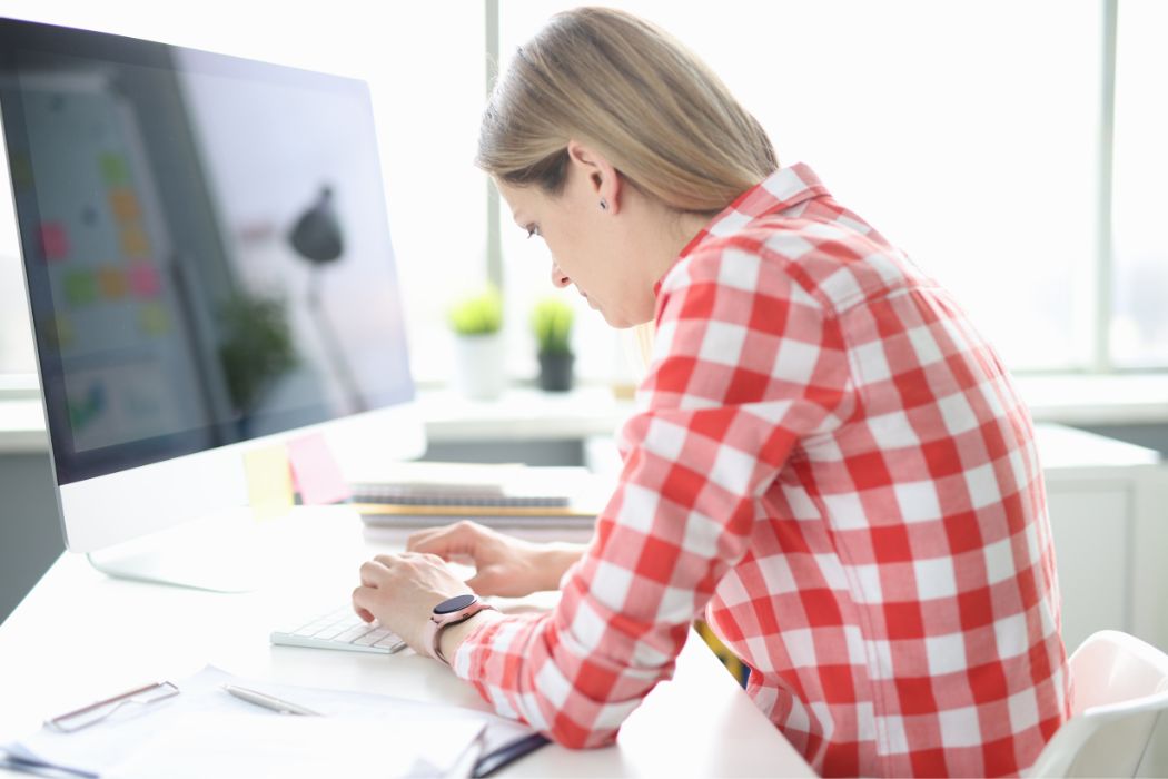 Žena sedí v kanceláři a soustředěně píše na bílou klávesnici. Je oblečená v červeno-bíle kostkované košili a má náramkové hodinky. Před ní je stůl s ležícími brýlemi a za ní je monitor počítače, na kterém jsou vidět barevné poznámky. Místnost je světlá s přirozeným světlem procházejícím skrze okno v pozadí.
