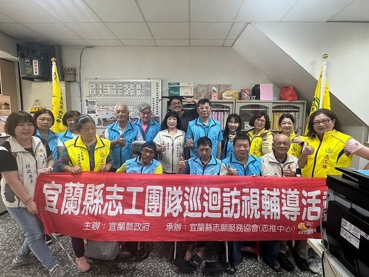 宜蘭縣政府志工團隊成員身穿黃色與藍色背心，手持「宜蘭縣志工團隊巡迴訪視輔導活動」紅色布條合影，眾人比出讚的手勢。