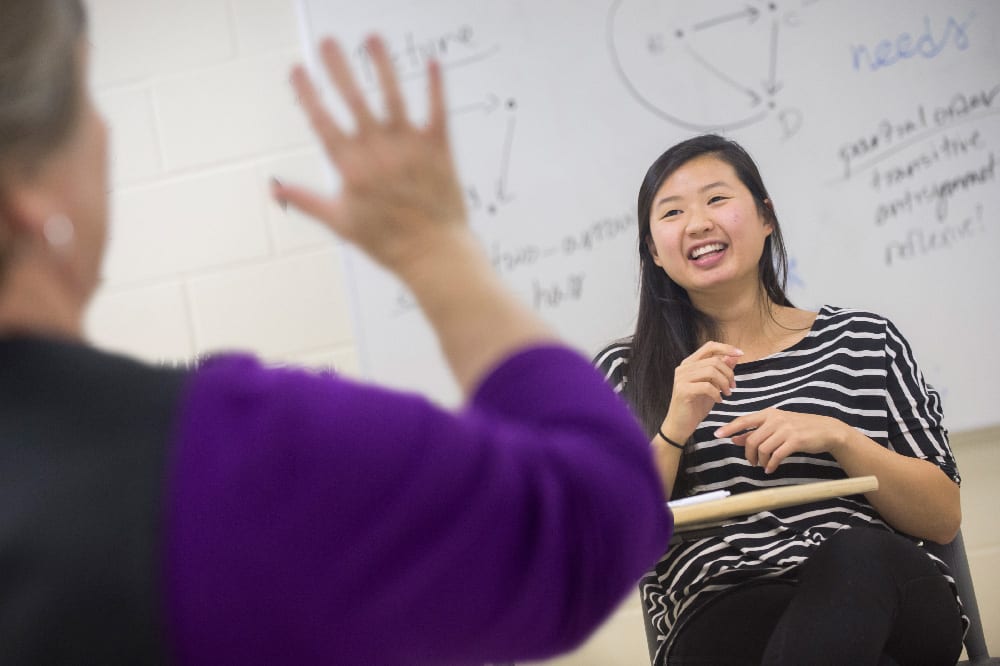 Student sits in front of a mathematical chart behind her, facing a blurry instructor, partially visible