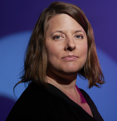 A white woman smiles gently at the camera standing in front of a vivid blue background, with a spotlight on her