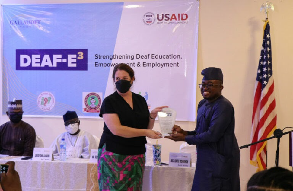 A white woman with dark pulled back hair is wearing a black short-sleeved shirt, a black mask and a green and pink patterned skirt while holding an award with Olufemi Ige. To her left, Olufemi is also holding the award, a black Nigerian man wearing glasses and a blue Senator dress. Behind them is an American flag, two men sitting at a table and a Gallaudet/USAID DEAF-E3 banner with the title 'Strengthening Deaf Education, Empowerment & Employment'. 