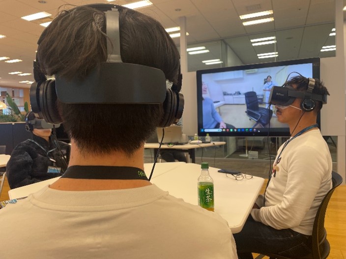 VRを装着した男性二人が椅子に座って体験している。会場スクリーンには参加者がVRで視聴している映像が流れている。