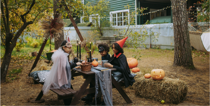 Barn utklädda i halloweendräkter. Sitter vid ett bord utomhus med pumpor runt omrking sig.