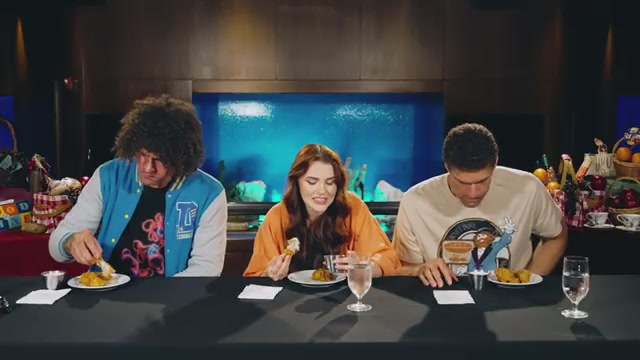 Three people are seated at a table, each with a plate of food, in front of a large blue aquarium.