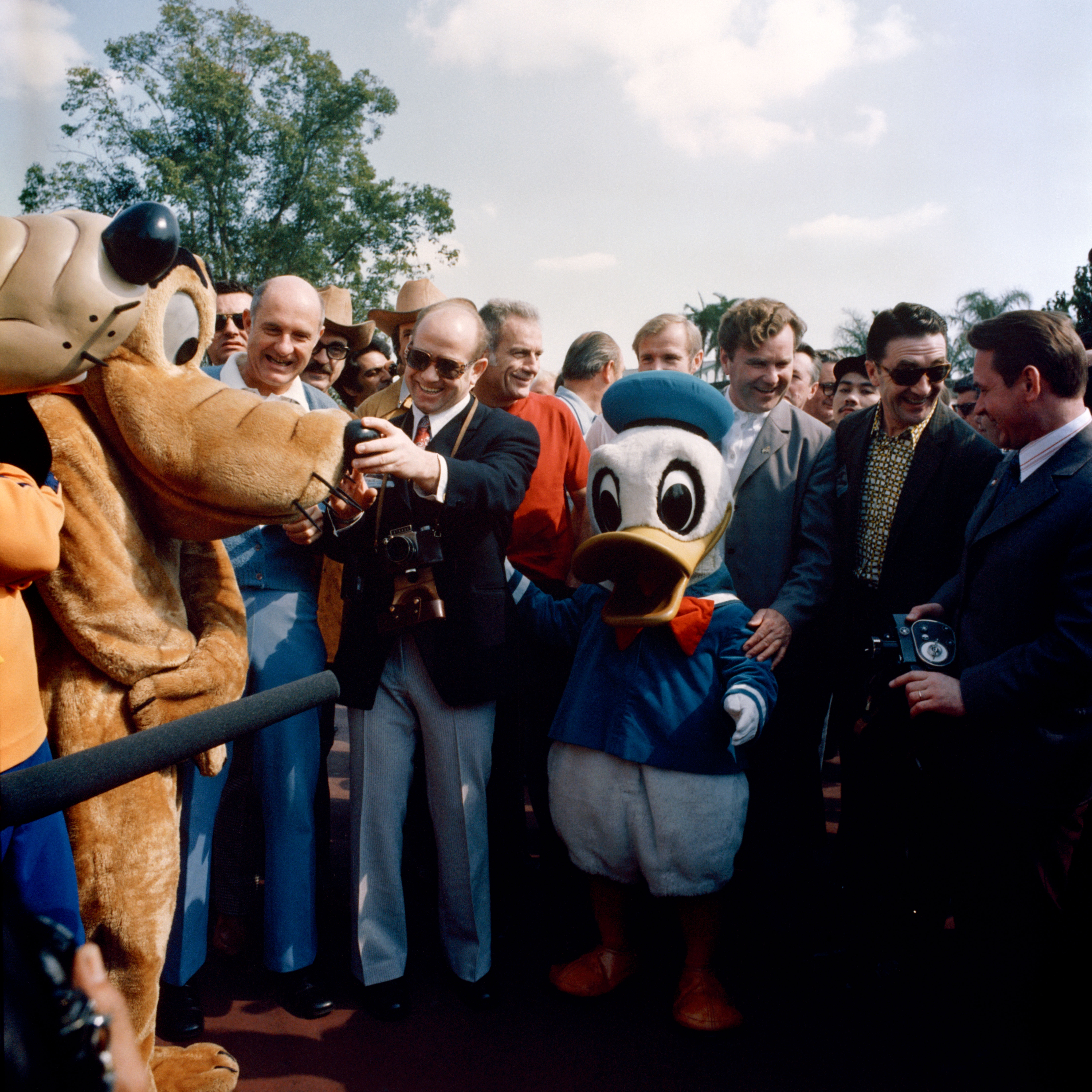 A man in a suit wearing sunglasses smiles while holding a camera, with two Disney characters, Pluto and Donald Duck, on either side of him, and other men in suits and casual attire behind them under a partially cloudy sky and trees.