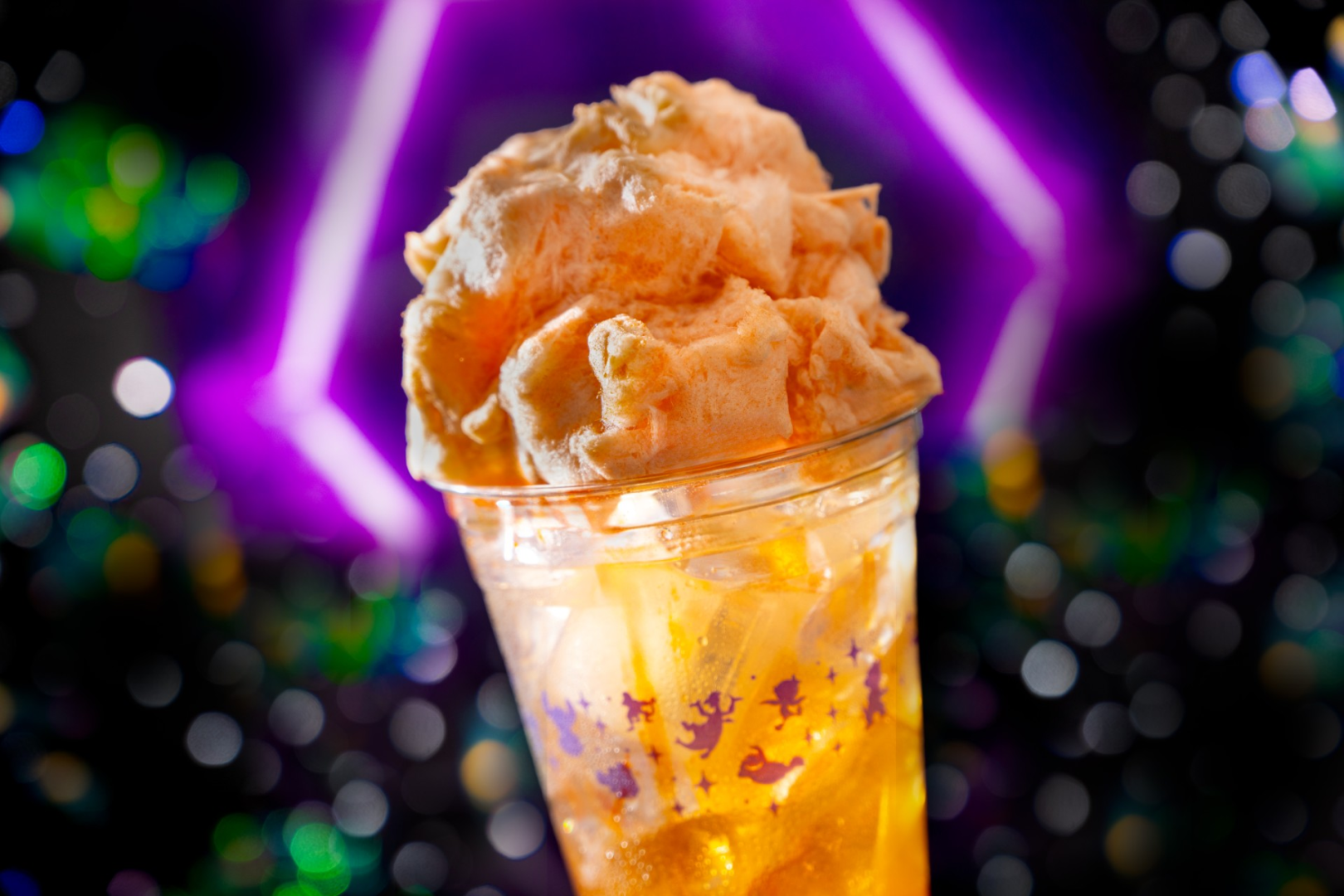 A drink in a clear printed cup topped with a large mound of orange cotton candy against a purple neon background