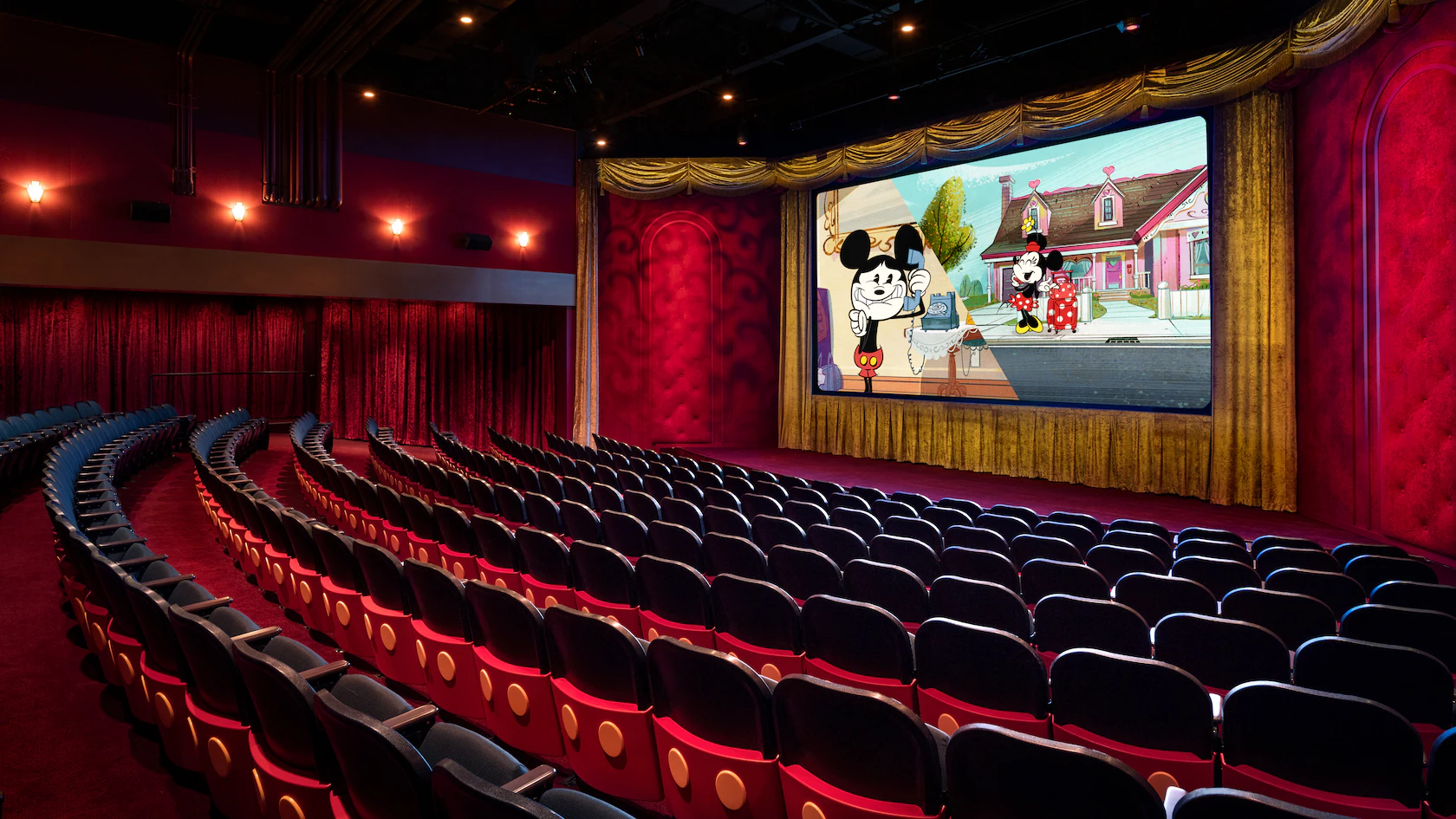 A movie theater with red and gold decor shows Mickey Mouse and Minnie Mouse on screen to rows of empty seats.
