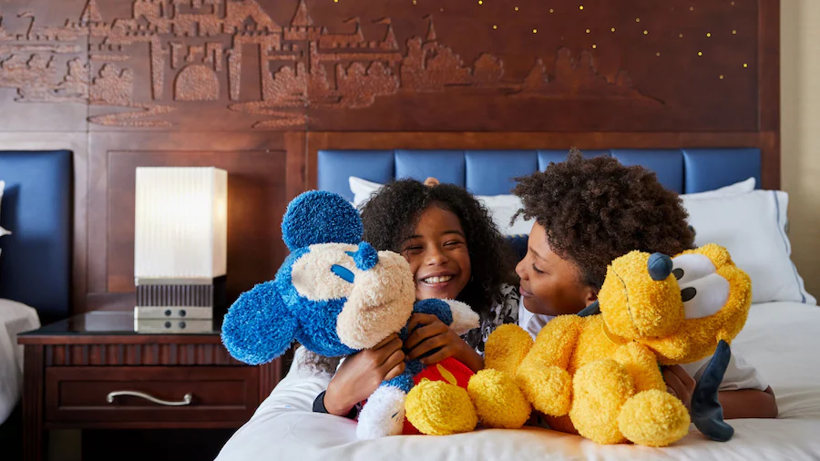 Two children smiling and cuddling Mickey Mouse and Pluto plush toys on a hotel bed at the Disneyland Hotel