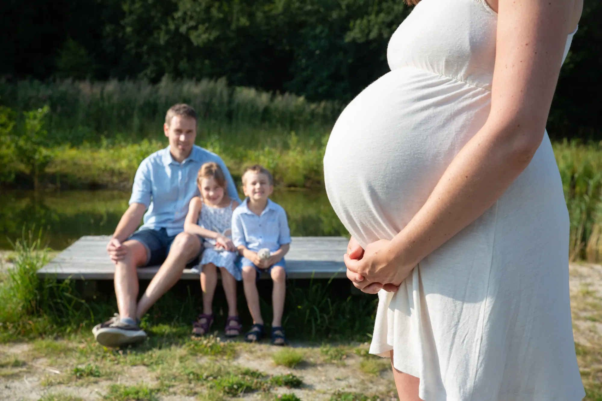 Ein Babybauch liebevoll mit den Händen umschlungen und im Hintergrund die erwartungsvollen Blicke der Familienmitglieder.  