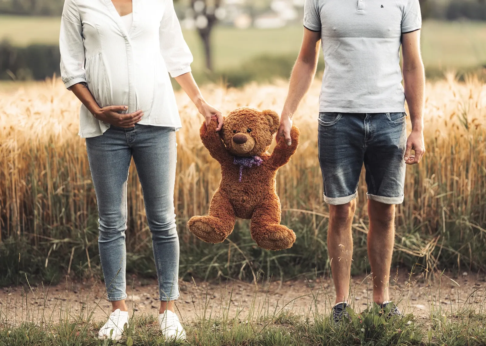 Eine Frau mit Babybauch und ein Mann halten einen ein Teddybär zwischen sich an den Händen.