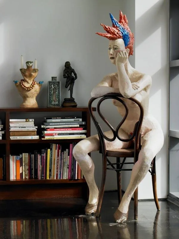 A seated pale figure on a bentwood chair in a room with books and sculpture.