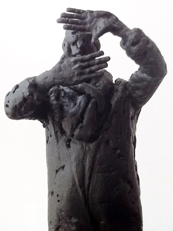A steel figure in a long coat, holding both hands up near its head.