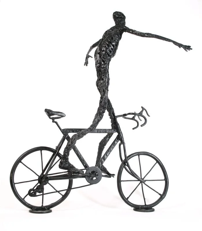 A steel figure standing on a bicycle with both arms outstretched.