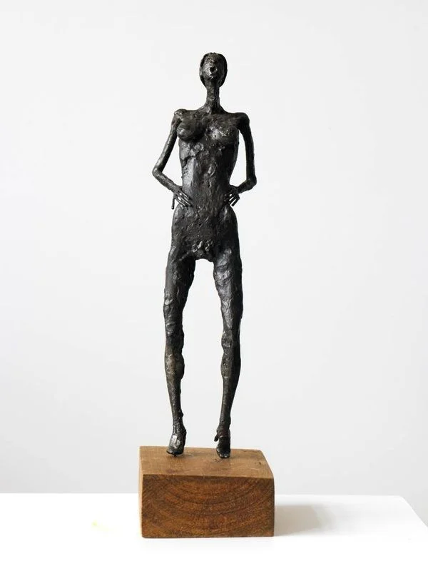 A narrow steel figure standing on a small wooden block with one hand raised.