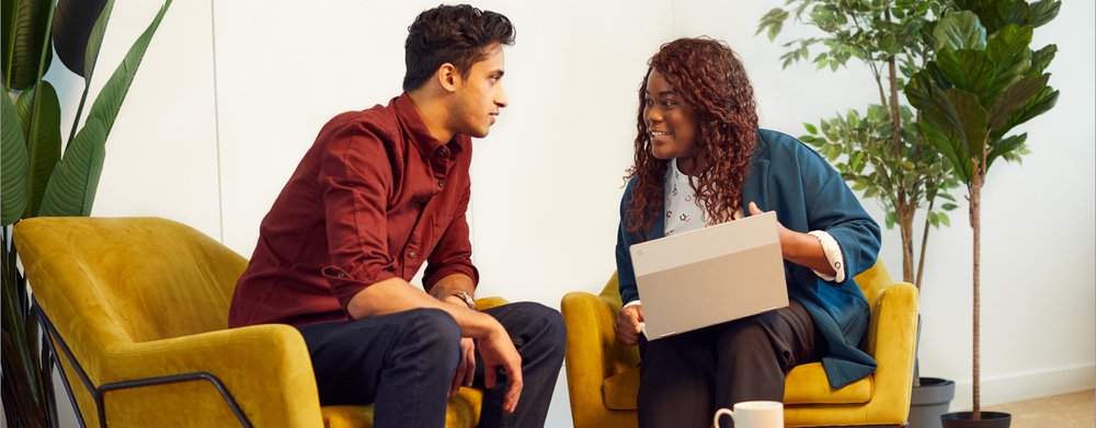 A man and woman are sat reviewing a laptop screen together, while the woman explains something.