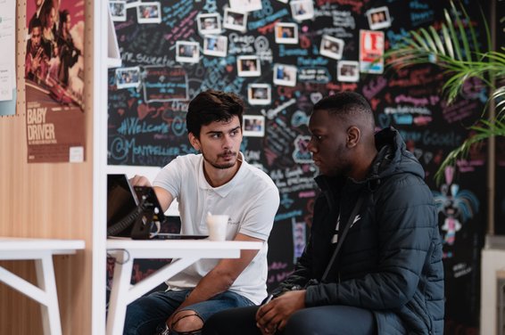 Zwei junge Männer sitzen auf Stühlen an einem Schreibtisch und schauen auf einen Computerbildschirm vor ihnen. Hinter ihnen ist eine Wand voller Brainstorming-Notizen und Fotos.