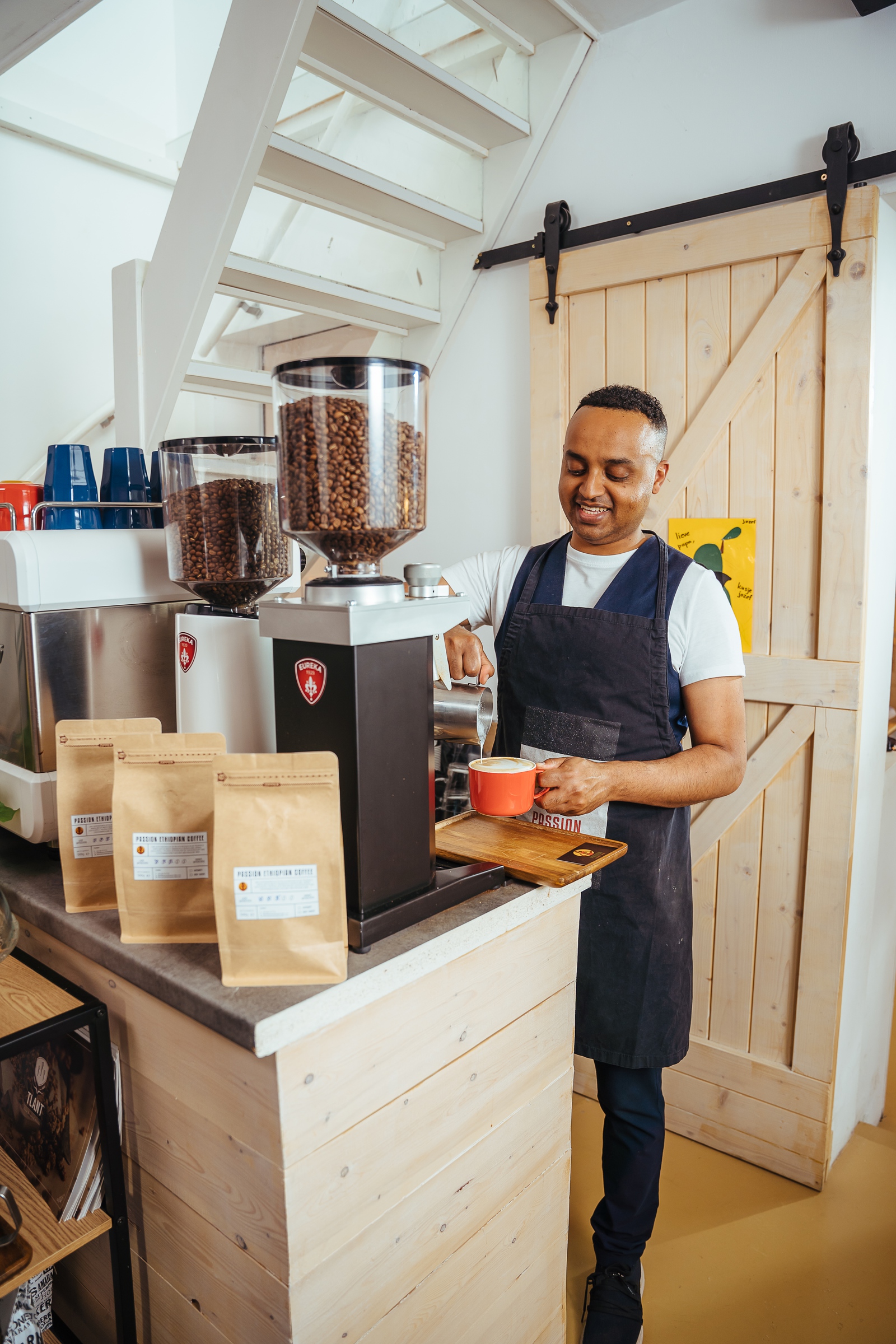 Een glimlachende man met een schort maakt koffie, op de tafel voor hem staat een professioneel koffiezetapparaat gevuld met koffiebonen en verschillende pakken koffie.