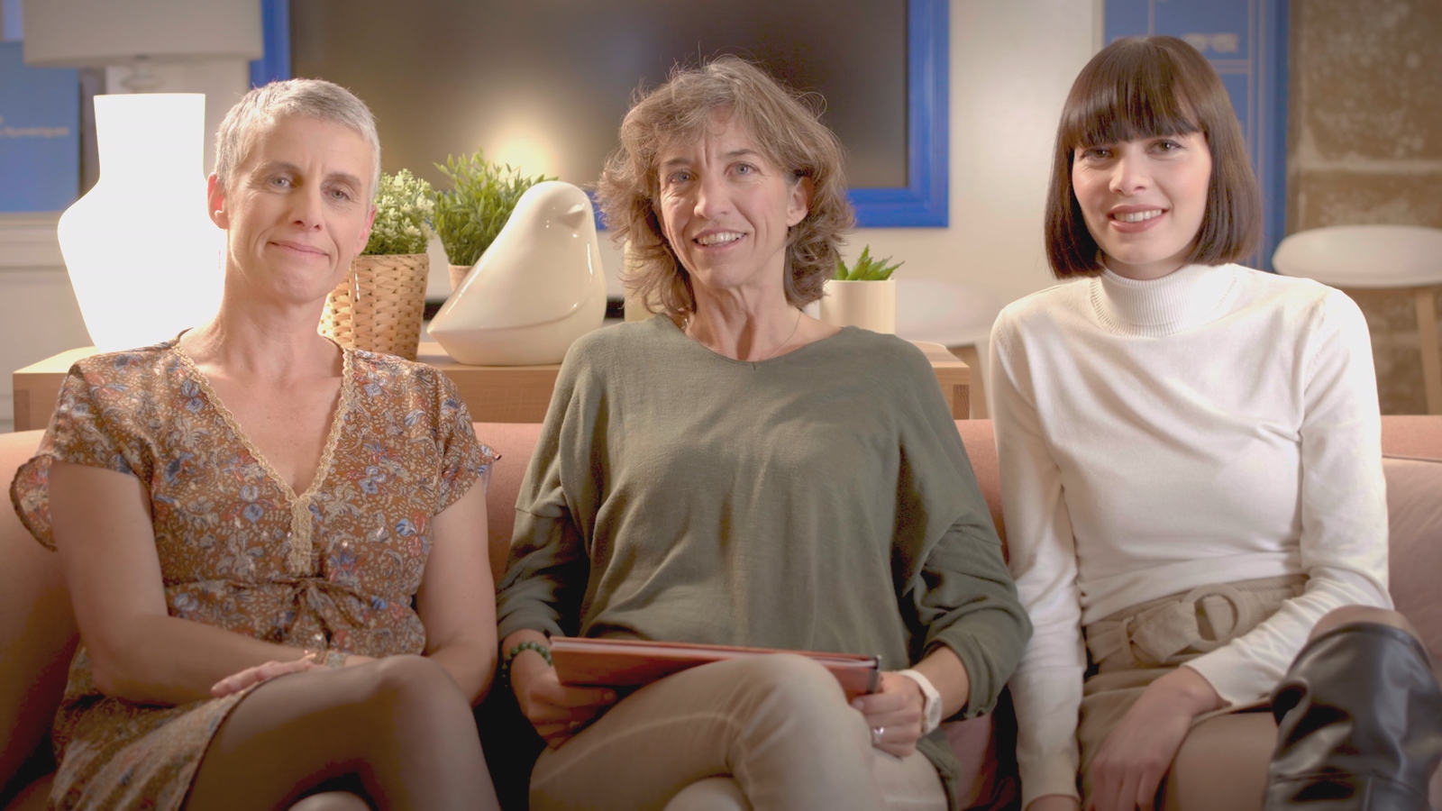 Trys moterys šypsosi į fotoaparatą sėdėdamos ant sofos.