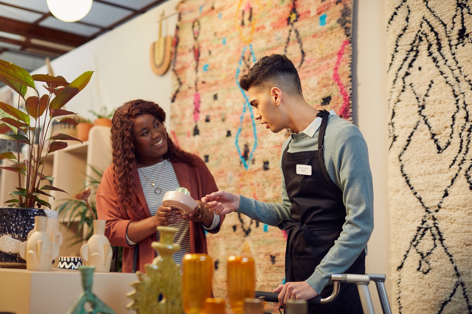 A smiling shopper in a store full of rugs, plants and ceramic ornaments asks a sales assistant in overalls about a product he is selling.