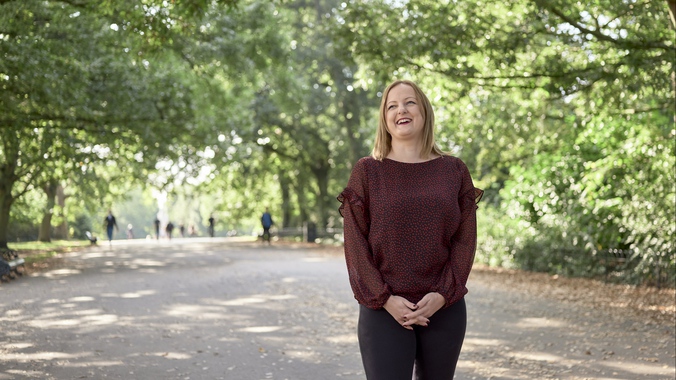 A woman smiles in a park, in front of a backdrop of trees, with dappled light shining through.